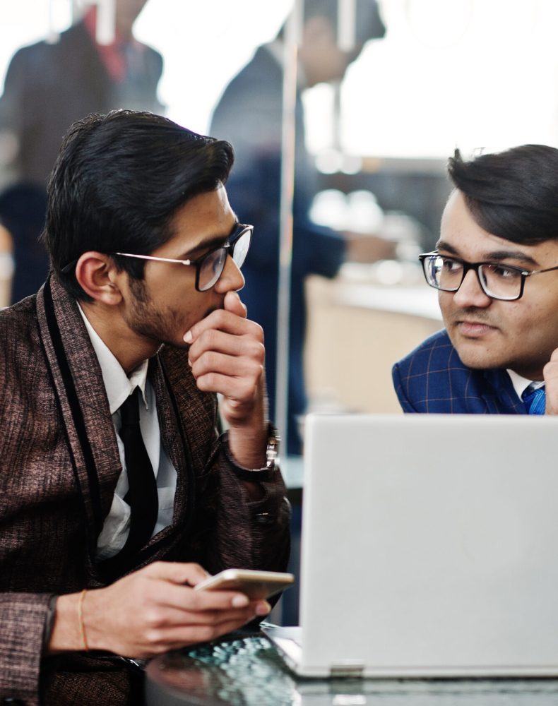 Two indian business man in suits sitting at office on cafe and looking at laptop.